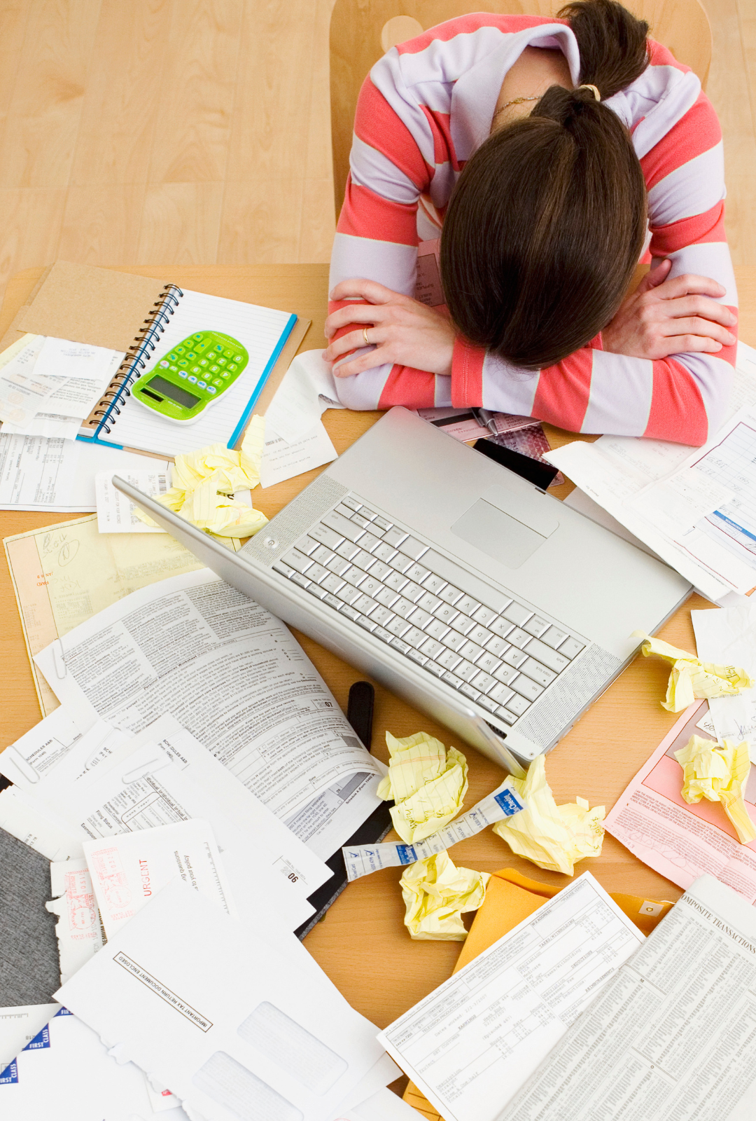 the image is of a lady with her head resting on her arms on a table surrounded by her laptops lots of paperworks and screwed up paper